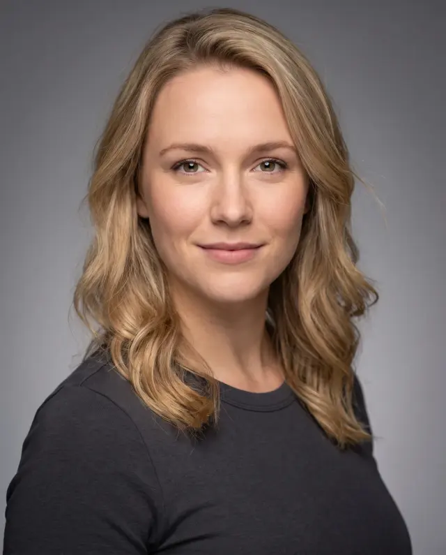 Actor headshot of a woman with wavy blonde hair in a charcoal top on a grey backdrop