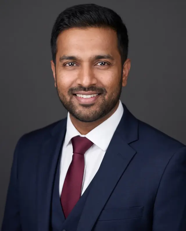 Corporate headshot of a bearded man in a navy three-piece suit and burgundy tie on a charcoal backdrop