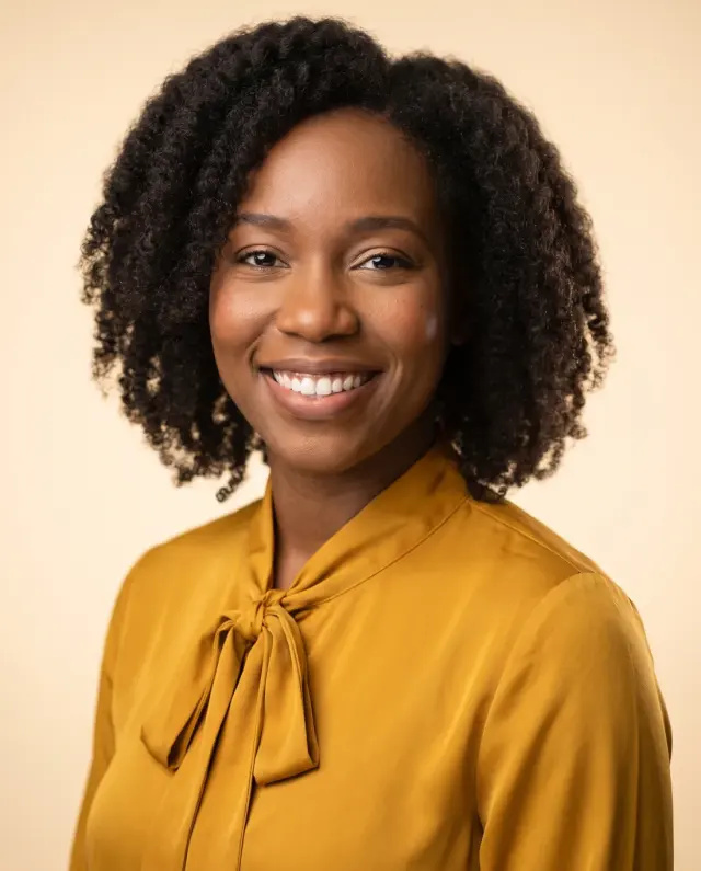 Professional headshot of a woman with curly hair in a mustard tie-neck blouse on a cream backdrop