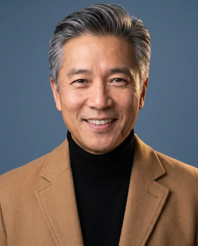 Executive headshot of a grey-haired man in a camel blazer and black turtleneck on a blue backdrop