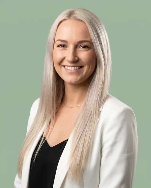 Professional headshot of a woman with long platinum-blonde hair in a white blazer on a sage-green backdrop