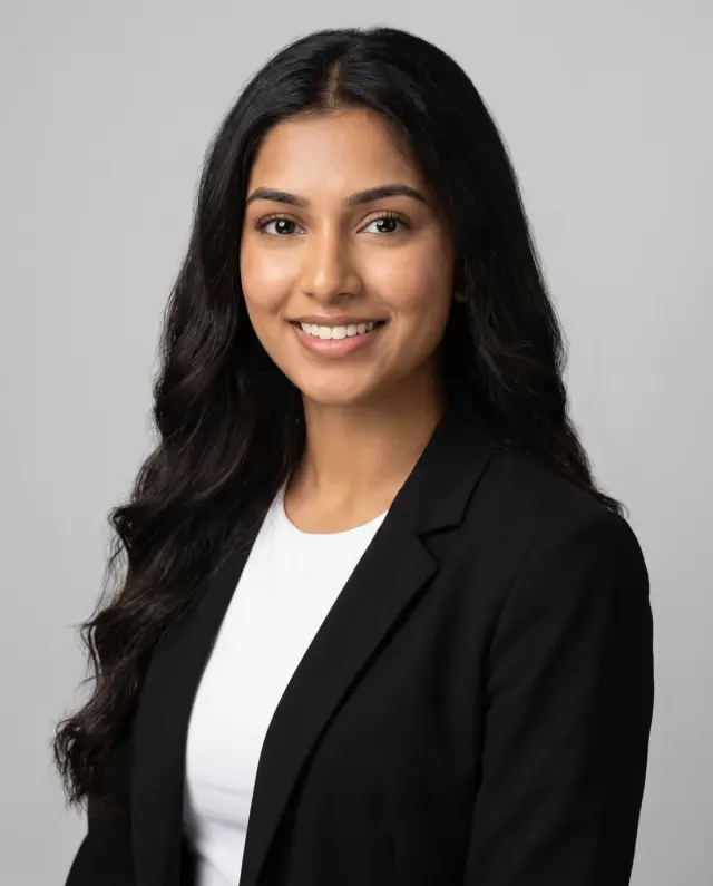 Professional headshot of a woman with long dark wavy hair in a black blazer on a light grey backdrop