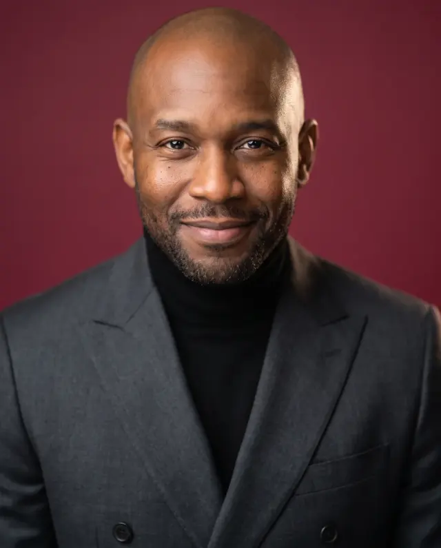 Executive headshot of a bald, bearded man in a charcoal jacket and black turtleneck on a deep red backdrop