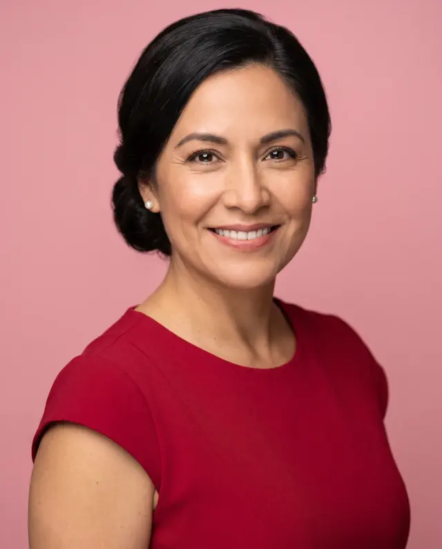 Professional headshot of a woman with her hair in a low bun, wearing a red dress on a pink backdrop