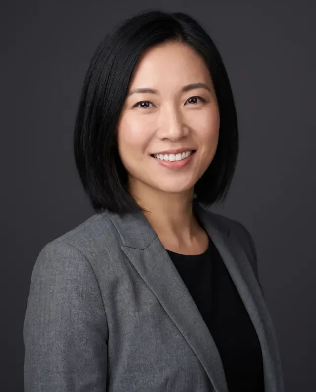 Professional headshot of a woman with a black bob in a grey blazer on a charcoal backdrop