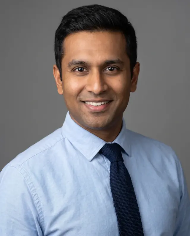 Corporate headshot of a man in a light-blue shirt and navy knit tie on a grey backdrop