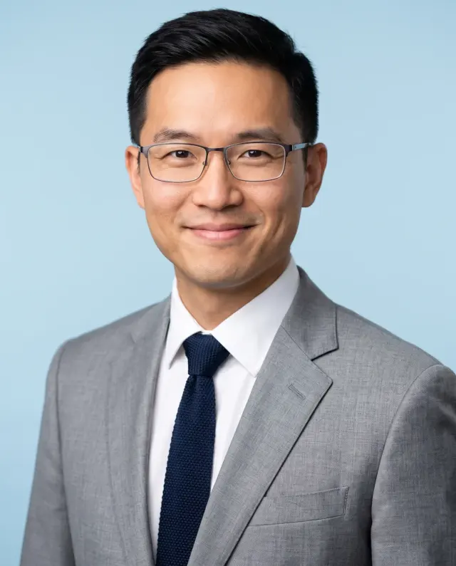 Corporate headshot of a man in glasses, a light grey suit and navy tie on a pale blue backdrop