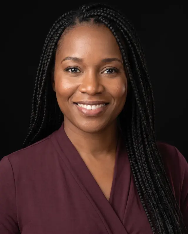 Professional headshot of a woman with long braids in a plum wrap top on a black backdrop