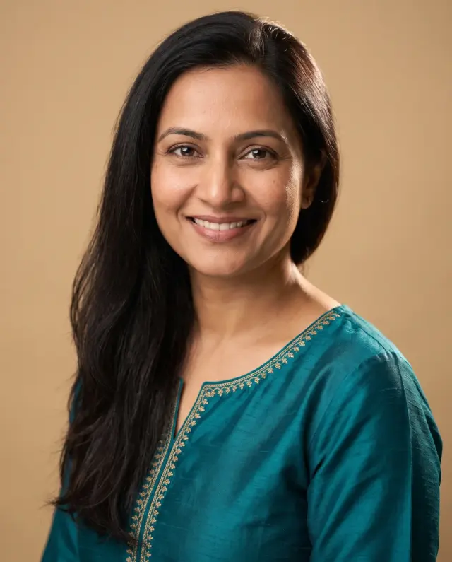 Professional headshot of a woman with long dark hair in a teal embroidered tunic on a tan backdrop
