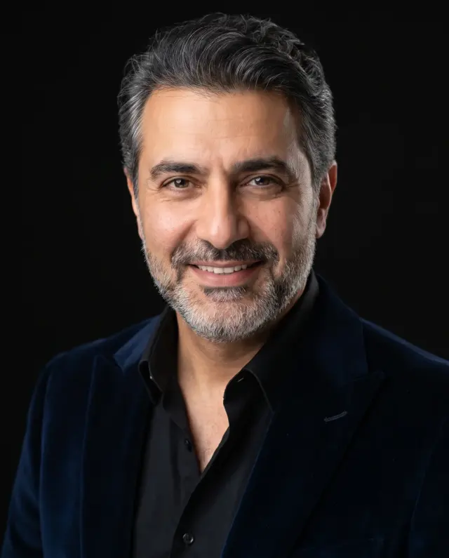 Executive headshot of a grey-bearded man in a navy velvet blazer and black shirt on a black backdrop