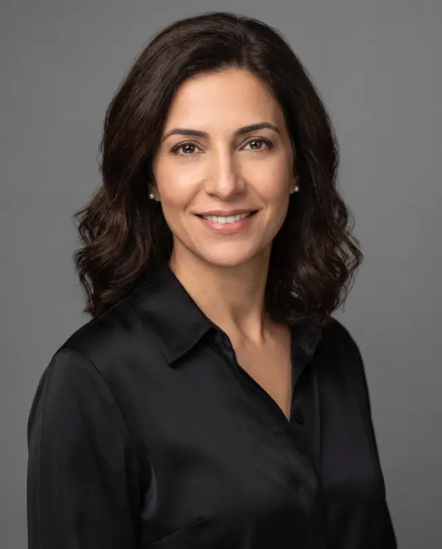Professional headshot of a woman with shoulder-length dark curls in a black satin shirt on a grey backdrop
