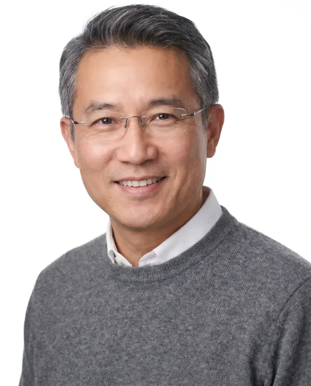 Professional headshot of a man in glasses and a grey sweater on a white backdrop