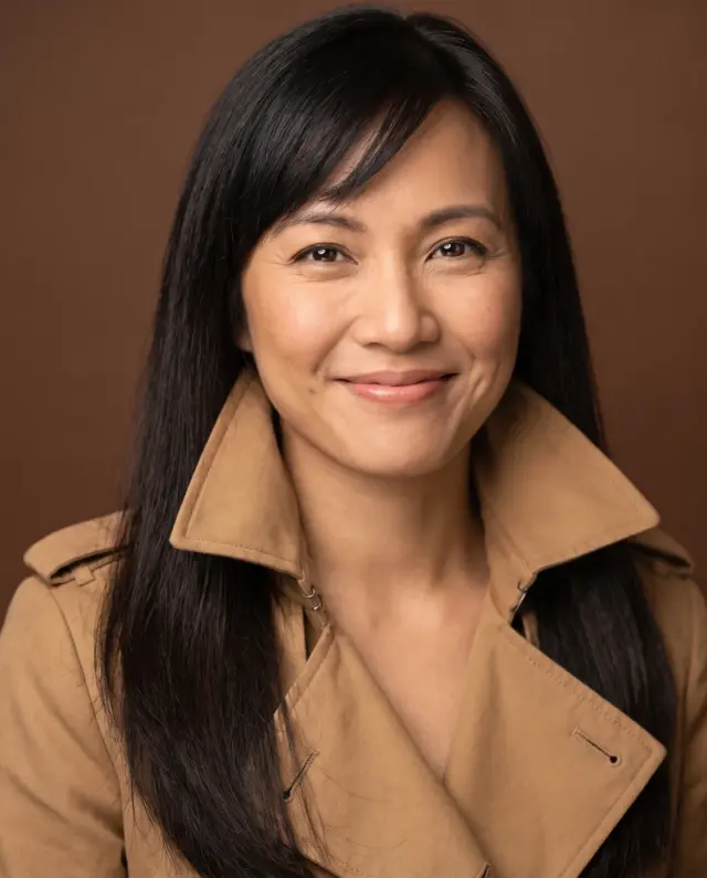 Actor headshot of a woman with long black hair and bangs in a camel trench coat on a brown backdrop