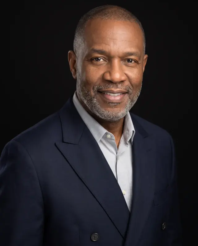 Executive headshot of a man with a grey beard in a navy double-breasted jacket on a black backdrop