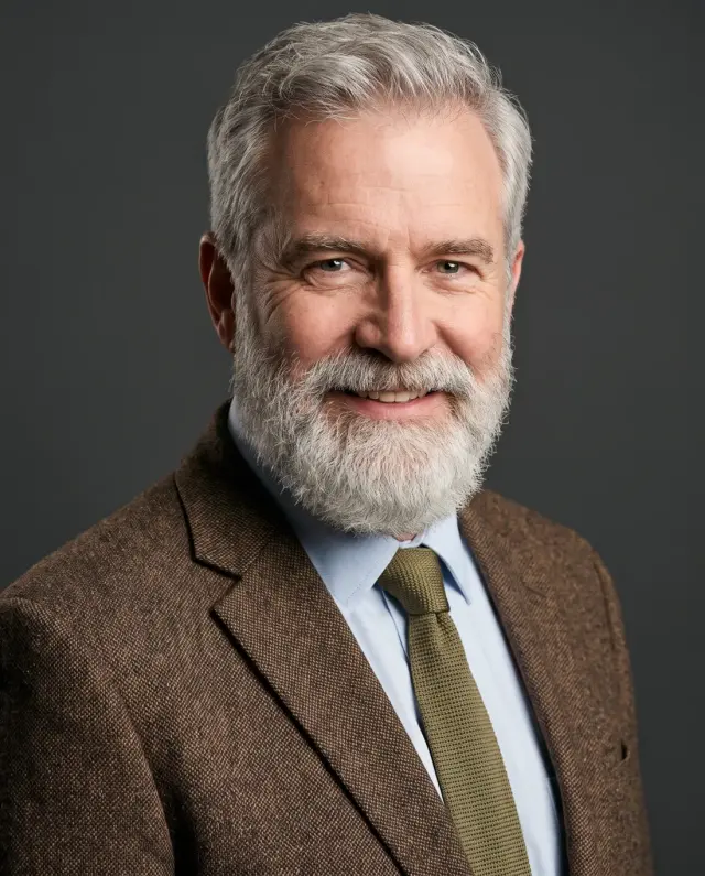 Executive headshot of an older man with a white beard in a tweed jacket and olive tie on a charcoal backdrop