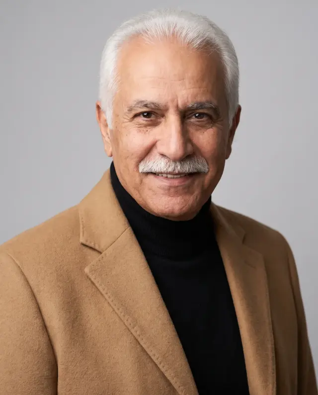 Executive headshot of an older man with a white moustache in a camel coat and black turtleneck on a grey backdrop