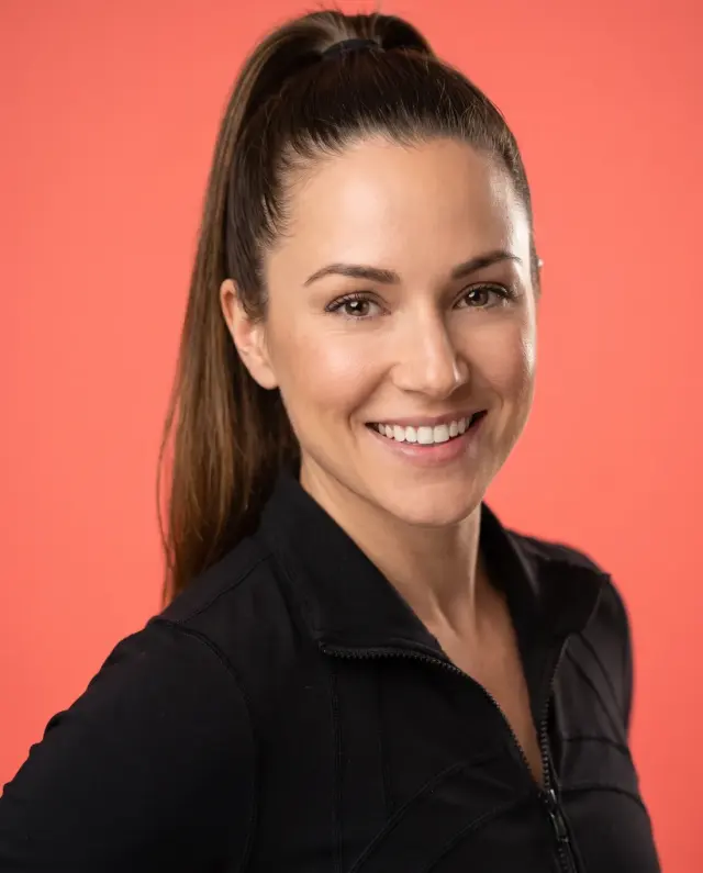 Actor headshot of a woman with a high ponytail in a black athletic jacket on a coral backdrop