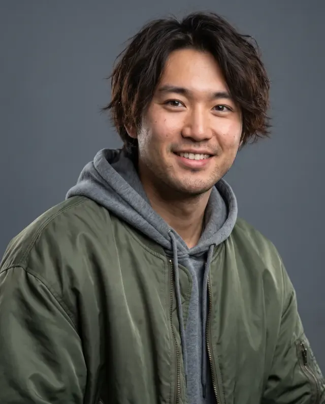 Actor headshot of a young man with tousled hair in a green bomber jacket and grey hoodie on a slate backdrop