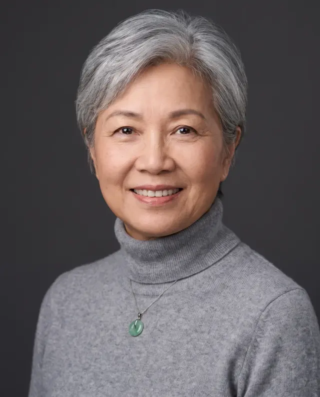 Headshot of an older woman with short silver hair in a grey turtleneck and jade pendant on a charcoal backdrop