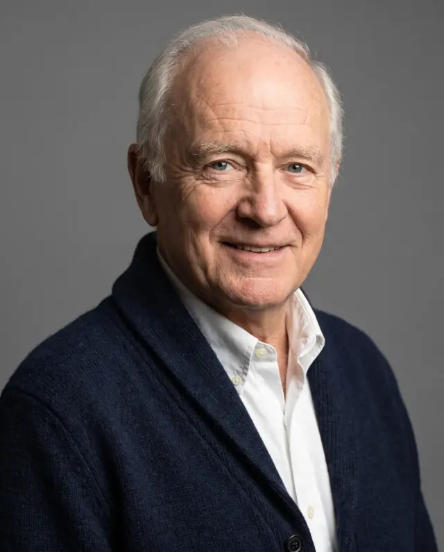 Headshot of an older man in a navy cardigan and white shirt on a grey backdrop