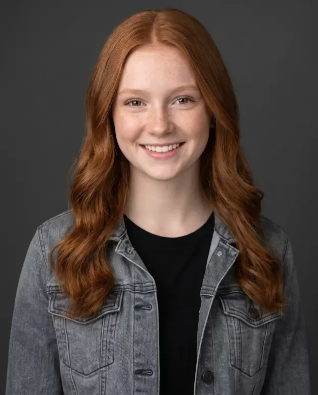 Young actor headshot of a teenage girl with long red hair in a grey denim jacket on a charcoal backdrop