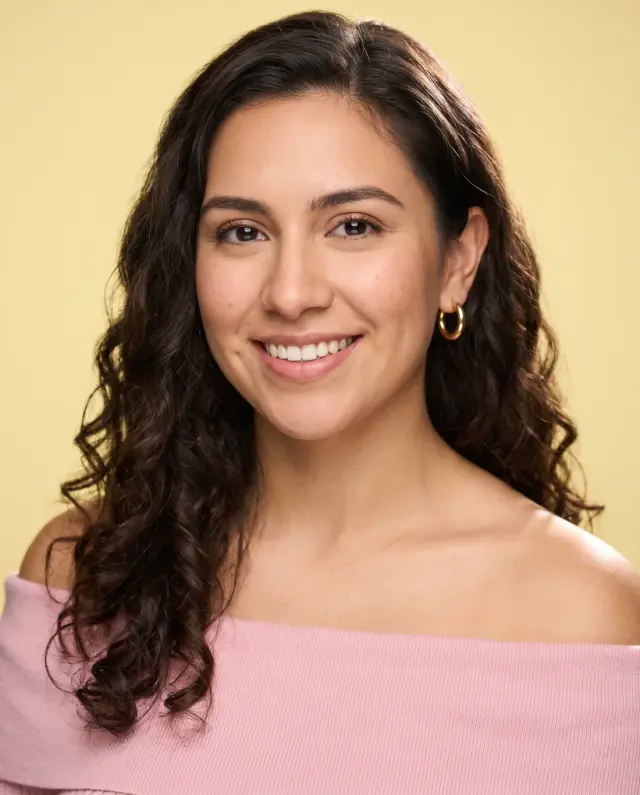Actor headshot of a woman with long dark curls in an off-shoulder pink top on a pale yellow backdrop